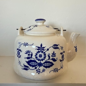 Pode incluir: Bule de cerâmica creme com padrões florais azuis. O bule tem um corpo arredondado, um bico curvo e uma tampa com um botão azul. O design floral inclui flores e folhas estilizadas. Um item de cozinha vintage.