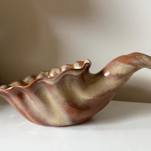 May include: A ceramic serving dish with a seashell design. The dish features a wavy rim and a curved handle. The colors are a mix of brown and beige, creating a marbled effect. This decorative piece is suitable for serving food or as a decorative item.