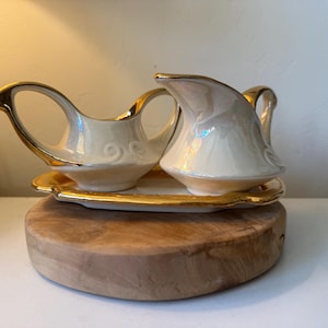 May include: A vintage cream-colored porcelain creamer and gravy boat set with gold trim, resting on a matching gold-trimmed tray. The set is displayed on a round wooden base, showcasing a classic design.