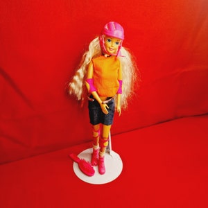 May include: A blonde doll wearing a pink helmet, orange shirt, blue shorts, and pink knee pads. The doll is standing on a white stand with a pink hairbrush on the side.