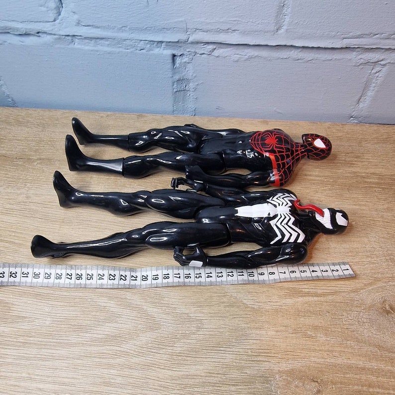 May include: Three black action figures of superheroes. The figures are posed in a standing position. The figure on the top has a red spider design on its chest. The figure in the middle has a white spider design on its chest. The figure on the bottom has a white skull design on its chest.