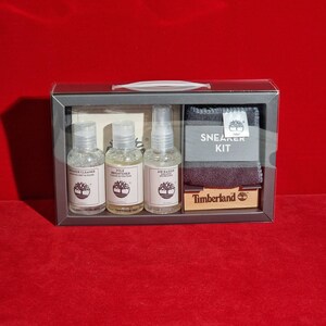 Op de afbeelding: Een grijze doos met een Timberland Sneaker Kit. De kit bevat drie flesjes reinigingsoplossing: Sneaker Cleaner, Sole Brightener en Air Raider Sneaker Deodorizer. De kit bevat ook een grijze doek en een klein houten blokje met het Timberland logo.