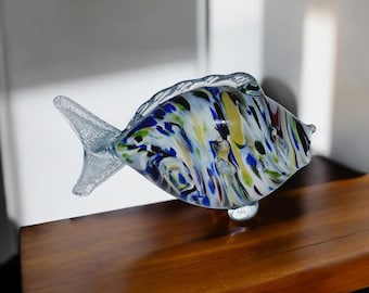 May include: A glass fish figurine with a clear body and a colorful, swirling pattern of blue, green, yellow, and white. The fish is standing on a wooden surface.