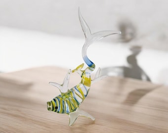 May include: A glass figurine of a yellow and blue striped fish with a clear tail fin. The fish is posed in a jumping position with its tail raised high in the air.