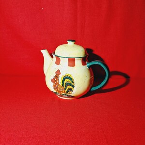 May include: A cream-coloured ceramic teapot with a green handle and a rooster design. The rooster is red, yellow, and green with a brown beak and eyes.