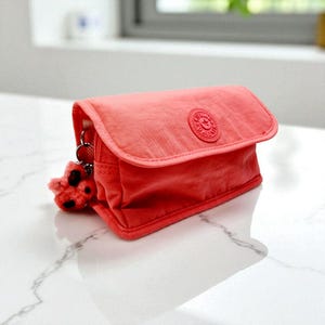 Peut inclure: Une pochette rectangulaire de couleur corail avec une fermeture à rabat. La pochette présente un logo circulaire sur le devant et un petit porte-clés flou avec des taches noires attaché à la fermeture éclair. Le sac est en matière lisse, possiblement en nylon.