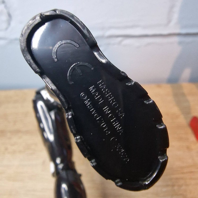 Pu&ograve; includere: Piede di action figure in plastica nera con il testo "HASBRO, INC. MADE IN CHINA, &copy; MARVEL 2014" sul fondo.