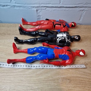 Pu&ograve; includere: Quattro action figure di supereroi, tra cui Spider-Man, Venom e due versioni di Spider-Man in costume rosso e nero. Le figure sono posizionate su una superficie di legno con un metro a nastro.