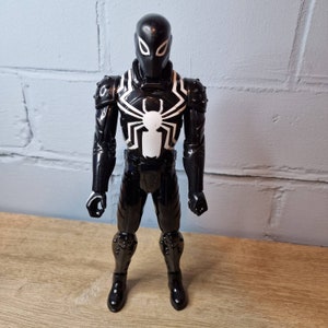 Pu&ograve; includere: Figurina d'azione in bianco e nero di un supereroe che indossa una tuta nera con un simbolo di ragno bianco sul petto. La figura ha un casco nero con occhi bianchi.