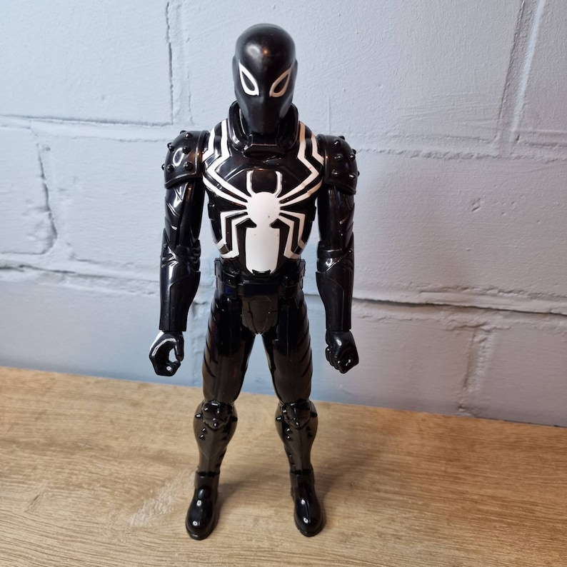 Pu&ograve; includere: Figurina d'azione in bianco e nero di un supereroe che indossa una tuta nera con un simbolo di ragno bianco sul petto. La figura ha un casco nero con occhi bianchi.