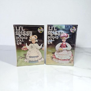 Pode incluir: Duas caixas vintage "Lil Missy Beaded Doll Kit". As caixas são pretas com a imagem e o texto da boneca. Uma boneca usa um vestido branco, a outra um vestido vermelho. O texto nas caixas diz "Lil Missy Beaded Doll Kit".