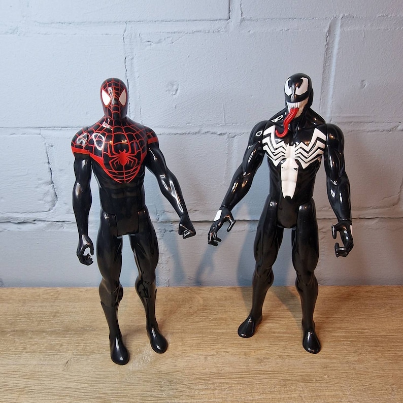 May include: Two action figures, one black and red Spider-Man and one black Venom, stand on a wooden surface. Both figures have articulated limbs and are posed in action stances.