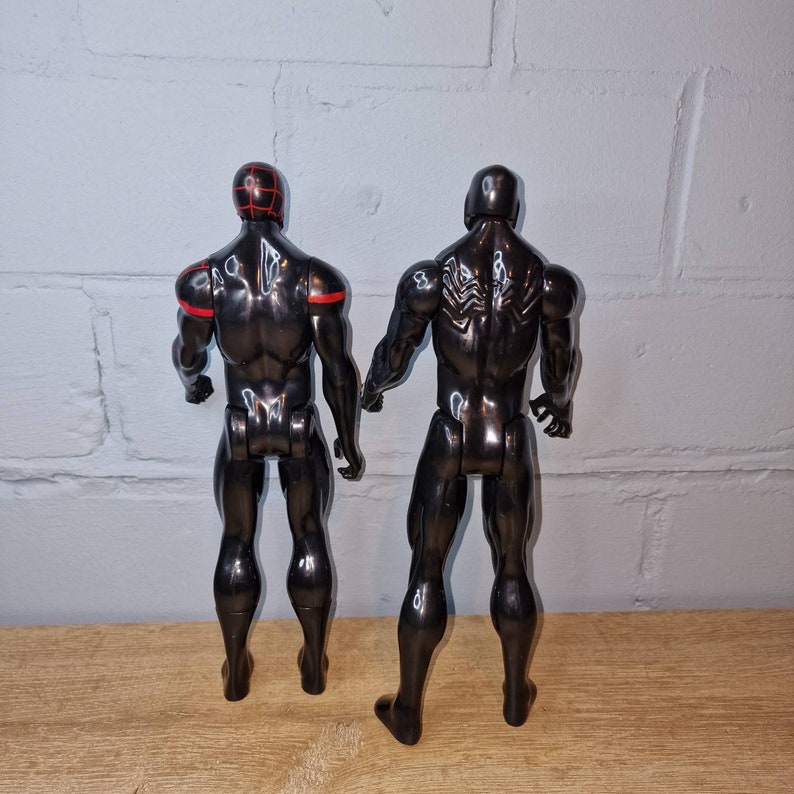 May include: Two black action figures, one with red accents and the other with a web design on the back. The figures are posed standing with their backs to the camera.