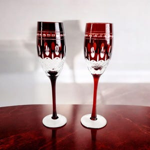 May include: Two red wine glasses with intricate cut crystal designs. The glasses have a clear base and a deep red stem.