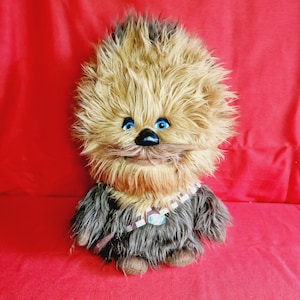 May include: A brown and white furry Chewbacca plush toy with blue eyes. The toy is wearing a brown vest with a black strap.