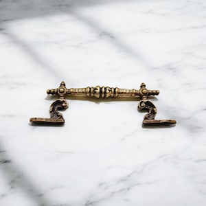 Pode incluir: Uma puxador de gaveta de metal dourado com detalhes ornamentados. O puxador tem uma barra central com um padrão decorativo e duas extremidades curvas.