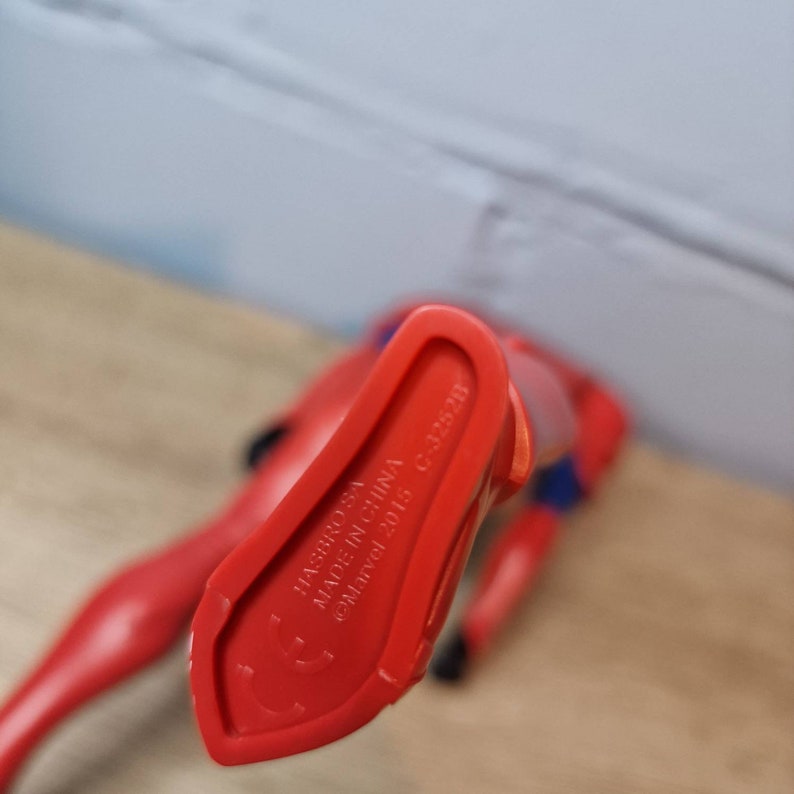 Pu&ograve; includere: Primo piano del fondo di una action figure di plastica rossa. Il piede della figura &egrave; visibile. La figura &egrave; etichettata "HASBRO, INC. MADE IN CHINA &copy;Marvel 2015 C-3253/H"
