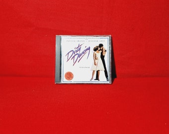 May include: A compact disc case with a red background and the title "Dirty Dancing" in white lettering. The case features a photo of Patrick Swayze and Jennifer Grey dancing.