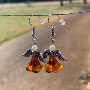 May include: A pair of angel-shaped earrings. The earrings feature silver-colored metal wings and a pearl bead above a translucent amber-colored bell-shaped body. The earrings hang from silver-colored hooks.