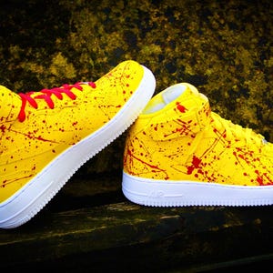 May include: Pair of yellow high-top sneakers with red splatter paint design and white soles. The sneakers have red laces and the word "AIR" is visible on the side of the sole. The shoes are on a dark wooden surface.