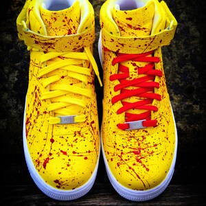 May include: Pair of yellow high-top sneakers with red paint splatter details. One shoe has yellow laces, the other has red laces. Both have white soles and a silver metal tag on the laces.
