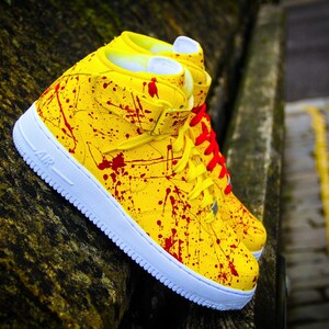 May include: A pair of high-top sneakers with a vibrant yellow base, splattered with red paint. The shoes feature white soles, yellow laces, and red laces on one shoe. The word "AIR" is visible on the side.