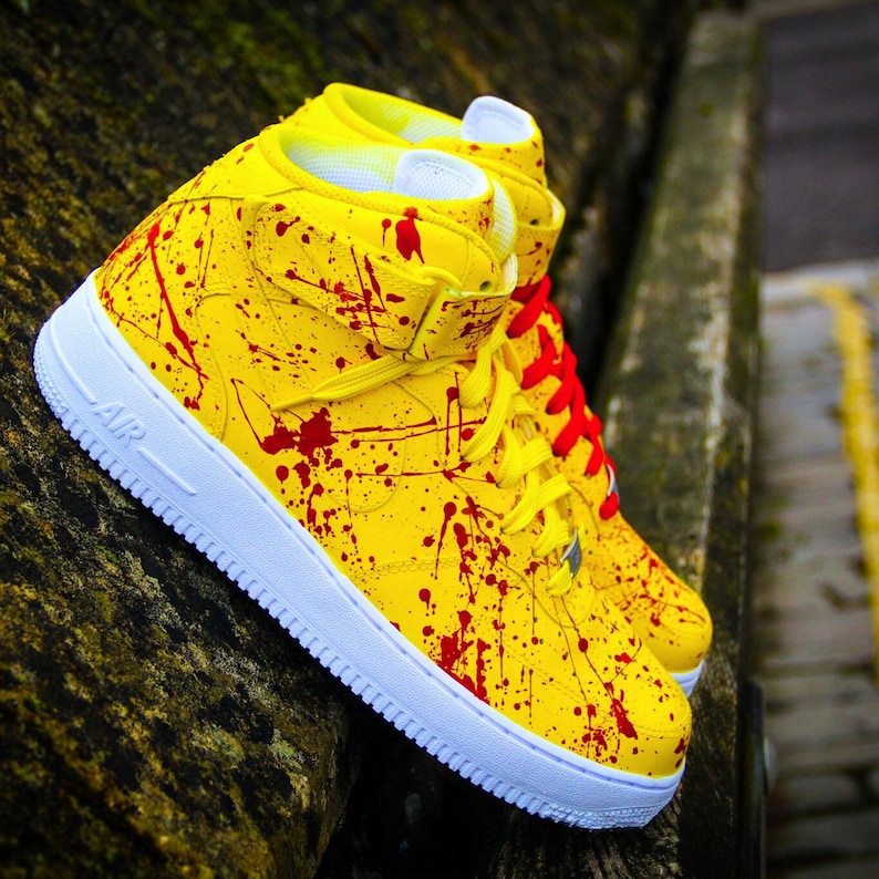May include: A pair of high-top sneakers with a vibrant yellow base, splattered with red paint. The shoes feature white soles, yellow laces, and red laces on one shoe. The word "AIR" is visible on the side.