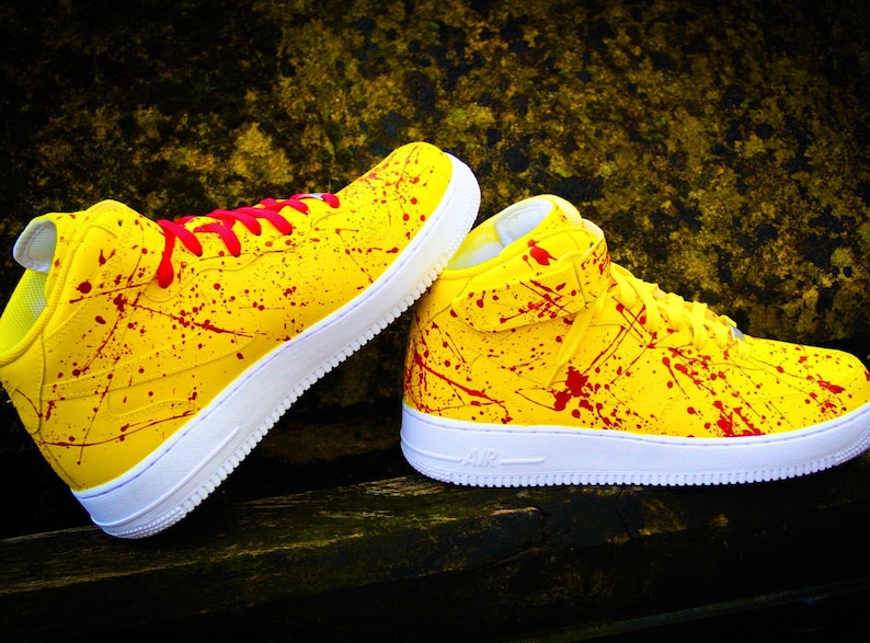 May include: Pair of yellow high-top sneakers with red splatter paint design and white soles. The sneakers have red laces and the word "AIR" is visible on the side of the sole. The shoes are on a dark wooden surface.