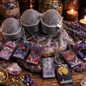 May include: A collection of tarot card pendants with silver chains, displayed with tea infusers, candles, and dried herbs. The pendants feature colorful illustrations and text, including "The Reader" and "A Call to Romance."