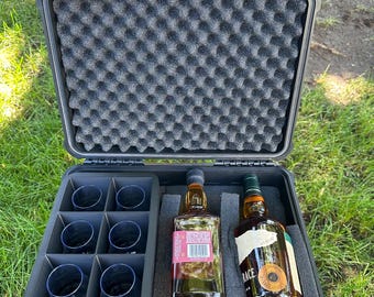 Puede incluir: Una maleta rígida negra con un interior de espuma. La maleta contiene dos botellas de whisky y seis vasos de chupito. La maleta tiene la palabra "Whiskey" impresa en la parte delantera.