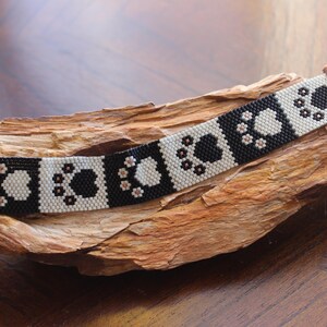 May include: A beaded bracelet featuring a repeating pattern of black and cream-colored paw prints. The bracelet has a copper-colored clasp and is displayed on a piece of wood. The paw print design is a charming detail.