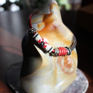 May include: A beaded bracelet with red, black, and white accents. The bracelet is displayed on a decorative stone sculpture with a marbled appearance. The bracelet has silver-toned metal accents and a leather band.
