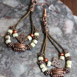 May include: Handmade copper earrings with a Celtic knot design. The earrings feature cream-colored beads, brown leather cords, and copper accents. The earrings are designed with a teardrop shape and are displayed on a stone surface.