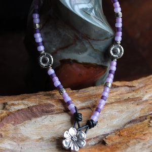 May include: A beaded necklace with a floral pendant. The necklace features small, light purple beads interspersed with darker purple beads and silver-toned metal accents. The pendant is a silver-colored flower.