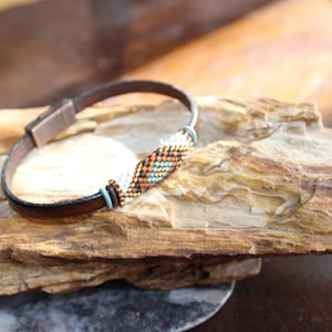 May include: A brown leather bracelet with a beaded center section. The beads are in a pattern of white, orange, and turquoise. The bracelet is resting on a piece of wood.