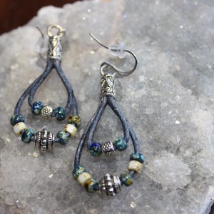 May include: A pair of handcrafted dangle earrings. Each earring features a teardrop-shaped design with a dark blue leather cord, adorned with silver-toned metal accents and colorful beads in shades of blue, white, and green. The earrings have a hook closure.