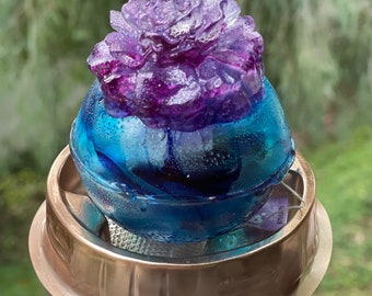 May include: A purple flower resin mold with a blue and white resin sphere inside. The mold is sitting on a copper-colored metal base.