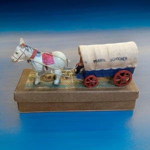 Pode incluir: Um cavalo de brinquedo branco puxando um vagão de brinquedo azul e branco com uma lona no topo que diz "Prairie Schooner". O vagão tem rodas vermelhas e o cavalo tem uma sela vermelha. O brinquedo está em uma caixa marrom.