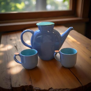 Może przedstawiać: Niebieski ceramiczny imbryk z pasującą pokrywką i dwoma filiżankami do herbaty. Imbryk i filiżanki mają jasnoniebieski wygląd zewnętrzny i turkusowe wnętrze. Filiżanki mają małe, okrągłe uchwyty w kolorze miedzi. Zestaw jest wystawiony na drewnianym stole.