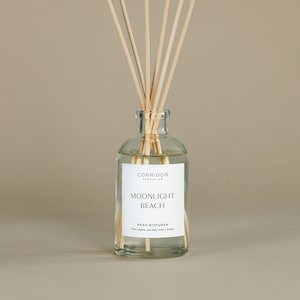 May include: A clear glass bottle with a white label that reads "CORRIDOR CANDLE CO. MOONLIGHT BEACH REED DIFFUSER Pear, agave, sea salt, rose + amber" and wooden sticks in the bottle.