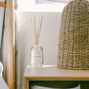 May include: A wooden nightstand with a white wicker basket, a clear glass bottle with a label that reads "CORRIDOR Torrey Pines Reed Diffuser", and a woven lamp base with a white lampshade.