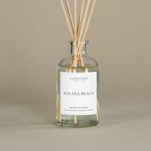 May include: A clear glass reed diffuser bottle with wooden reeds. The label reads "Solana Beach" and lists ingredients. The bottle is filled with a clear liquid. The background is a solid beige color.