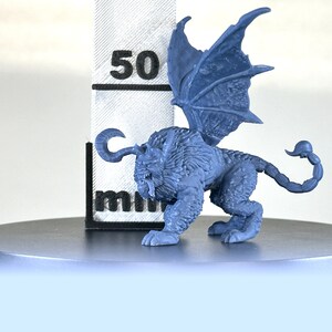 May include: A blue plastic miniature of a winged lion with a scorpion tail. The miniature is about 5 cm tall and is standing on a gray circular base.