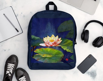 May include: A navy blue backpack featuring a watercolour design of a white and pink water lily on a green lily pad. The lily is surrounded by green leaves against a dark blue background.