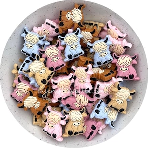 以下が含まれることがあります： ハイランド牛の形をしたカラフルなシリコンチャームが入ったボウル。チャームはピンク、ブルー、ブラウン、クリームの色合いです。