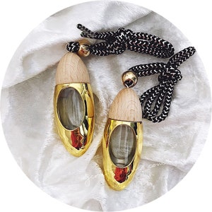 May include: Two gold-coloured, teardrop-shaped car air fresheners with wooden tops and clear glass windows. Each freshener has a black and white patterned cord with a gold bead. The fresheners are displayed on a white, textured surface.