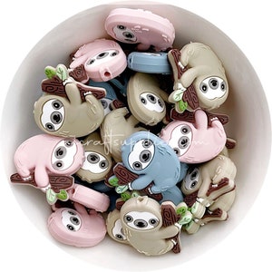 May include: A bowl filled with colourful silicone sloth charms. The charms are in shades of pink, brown, and blue. Each charm features a sloth hanging from a branch with leaves.