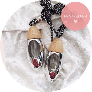 May include: Two silver car air fresheners with wooden tops and a black and white cord. The air fresheners are oval-shaped with a clear glass interior. A pink circle with the word "BESTSELLER" is in the background.