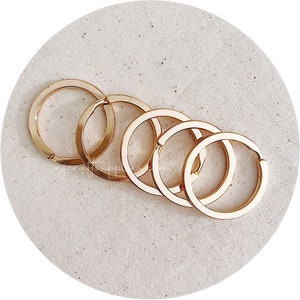 May include: Five gold-coloured metal key rings of varying sizes, arranged in a circle on a beige fabric surface.