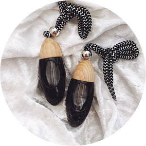 Op de afbeelding: Twee zwarte en houten autoluchtverfrissers met een zwart-wit koord dat er vanaf hangt. De luchtverfrissers liggen op een wit, pluche oppervlak.